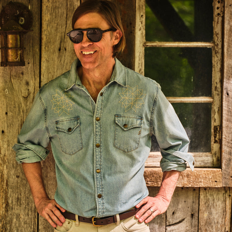 Man wearing sunglasses and a denim shirt standing in front of a wooden building.