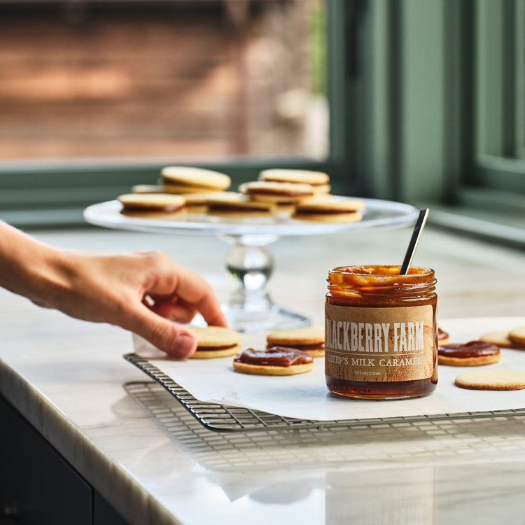 Person preparing cookies with a jar of 'Blackberry Farm' caramel on a kitchen counter.
