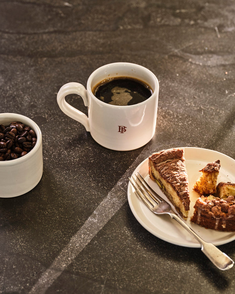 White mug with coffee, a small bowl of coffee beans, and a slice of cake on a dark surface.