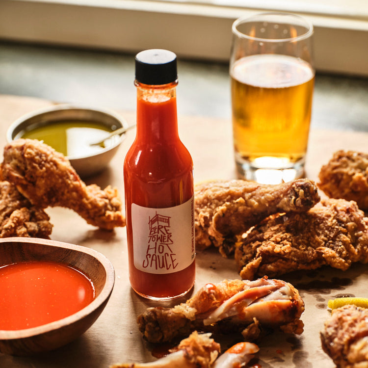 Bottle of hot sauce with fried chicken and a glass of beer on a table.