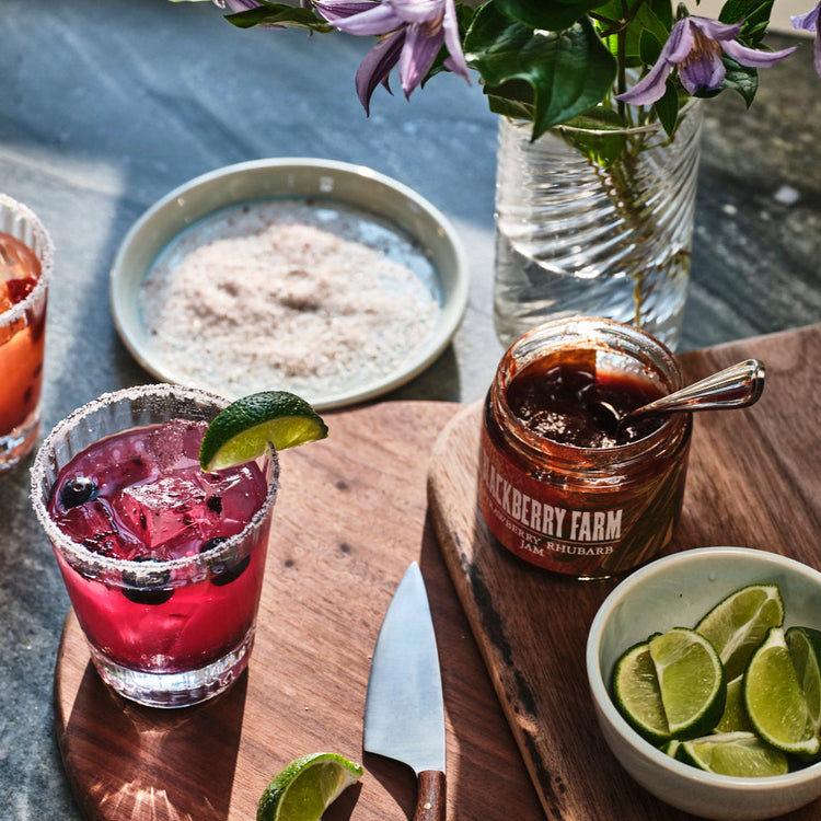 Two cocktails with garnishes on a wooden cutting board with a jar of jam and bowl of limes.