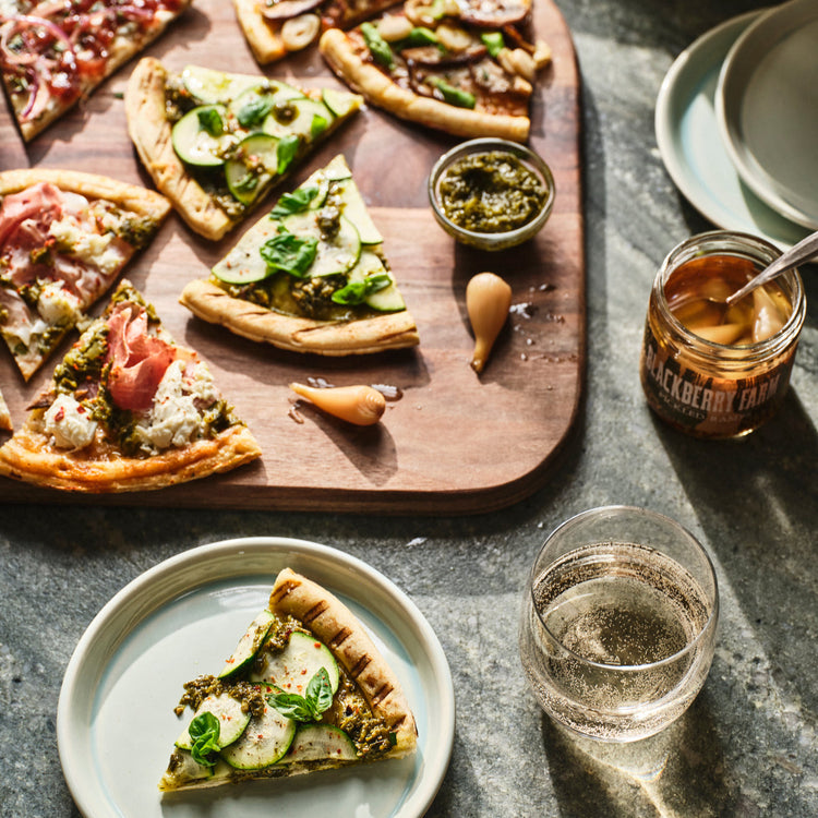 Wooden board with various pizzas and a slice on a plate, accompanied by a glass of water and a jar of honey.
