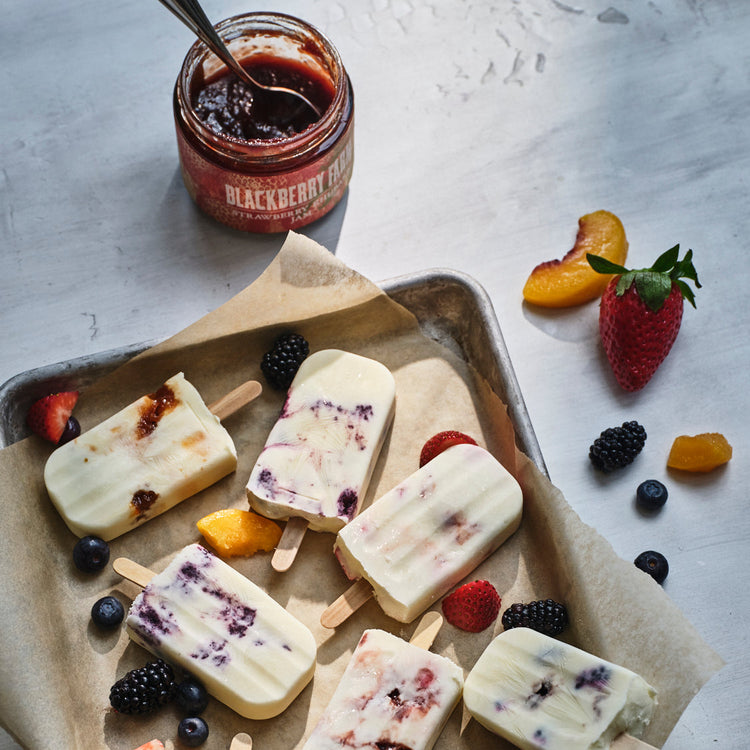 Popsicles with berries on a tray with a jar of blackberry jam in the background.