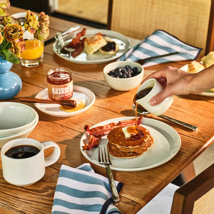 Person pouring syrup onto pancakes on a wooden table with breakfast items.