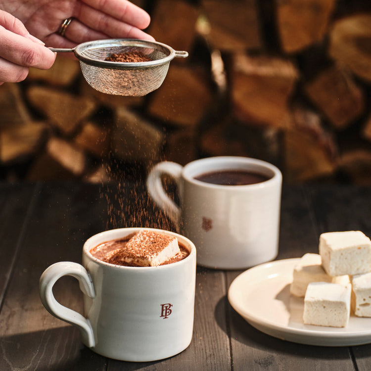 Person adding spices to a mug of hot chocolate with marshmallows on a wooden table.