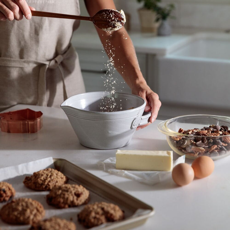 Person in a kitchen sifting flour into a bowl with baking ingredients on the counter.