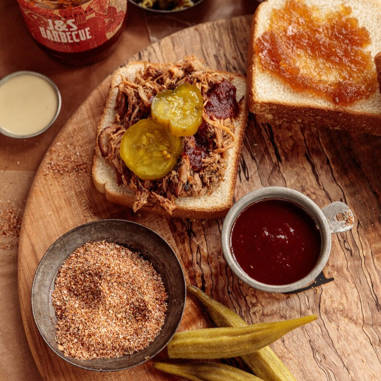 Wooden cutting board with barbecue sandwich, pickles, and various condiments on a wooden table.