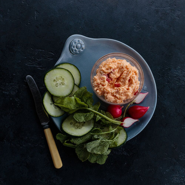 Plated dish with pimento cheese, sliced cucumbers, radishes, and greens on a dark surface.