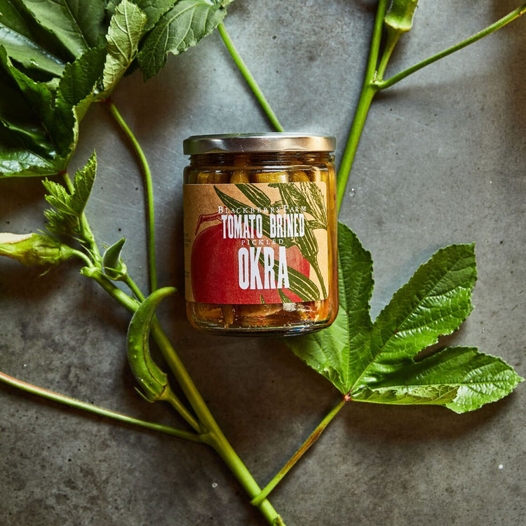 Jar of okra surrounded by green leaves on a gray surface