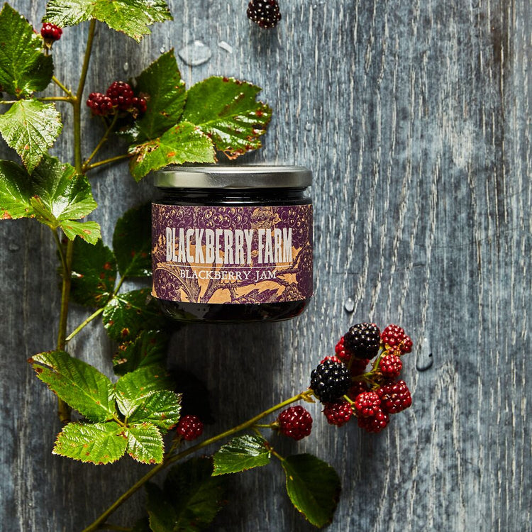 Jar of Blackberry Farm jam with blackberries and leaves on a wooden surface
