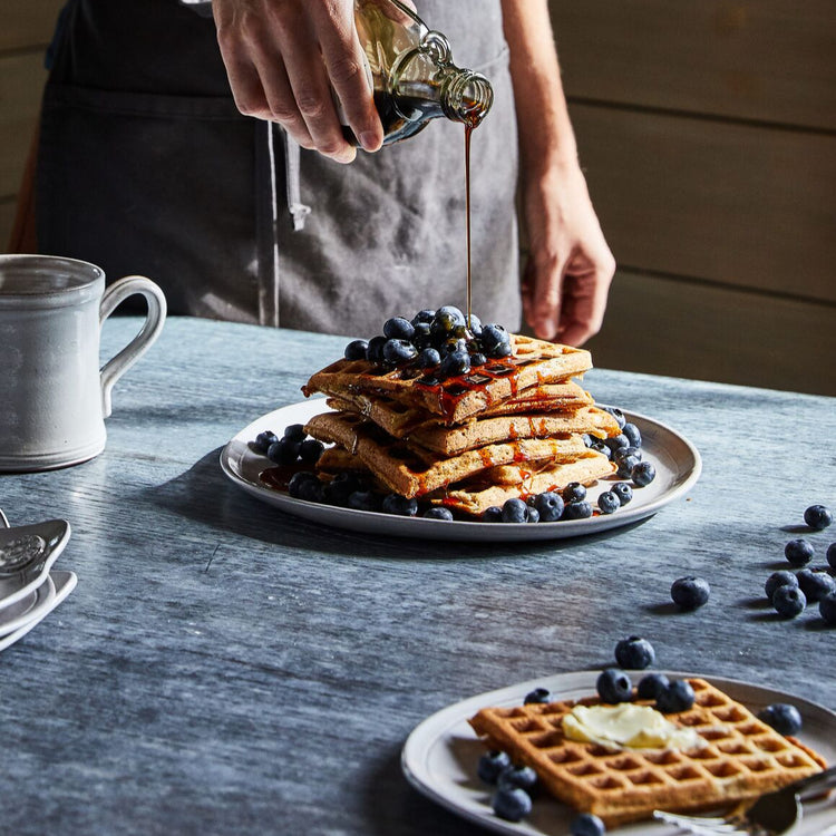 Person pouring syrup over waffles with blueberries on a wooden table.