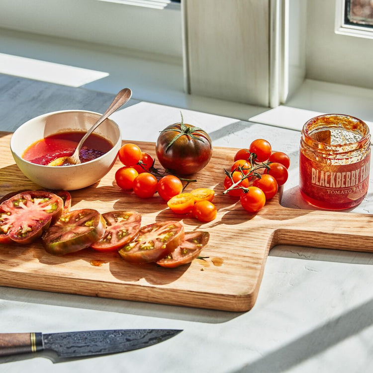 Wooden cutting board with sliced tomatoes, a bowl of sauce, and a jar of jam on a light-colored surface.