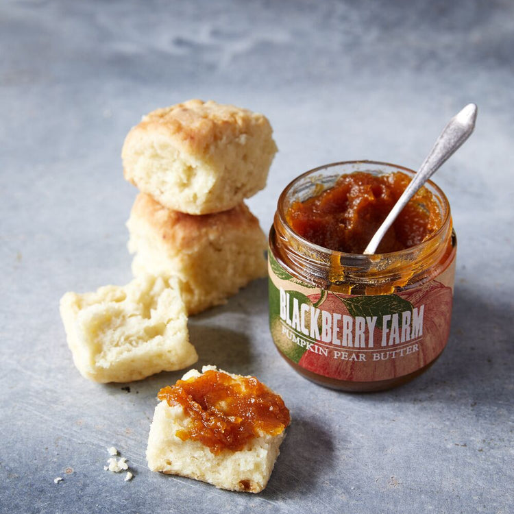 Jar of Blackberry Farm peach butter with a spoon, biscuit, and peach spread on a gray surface